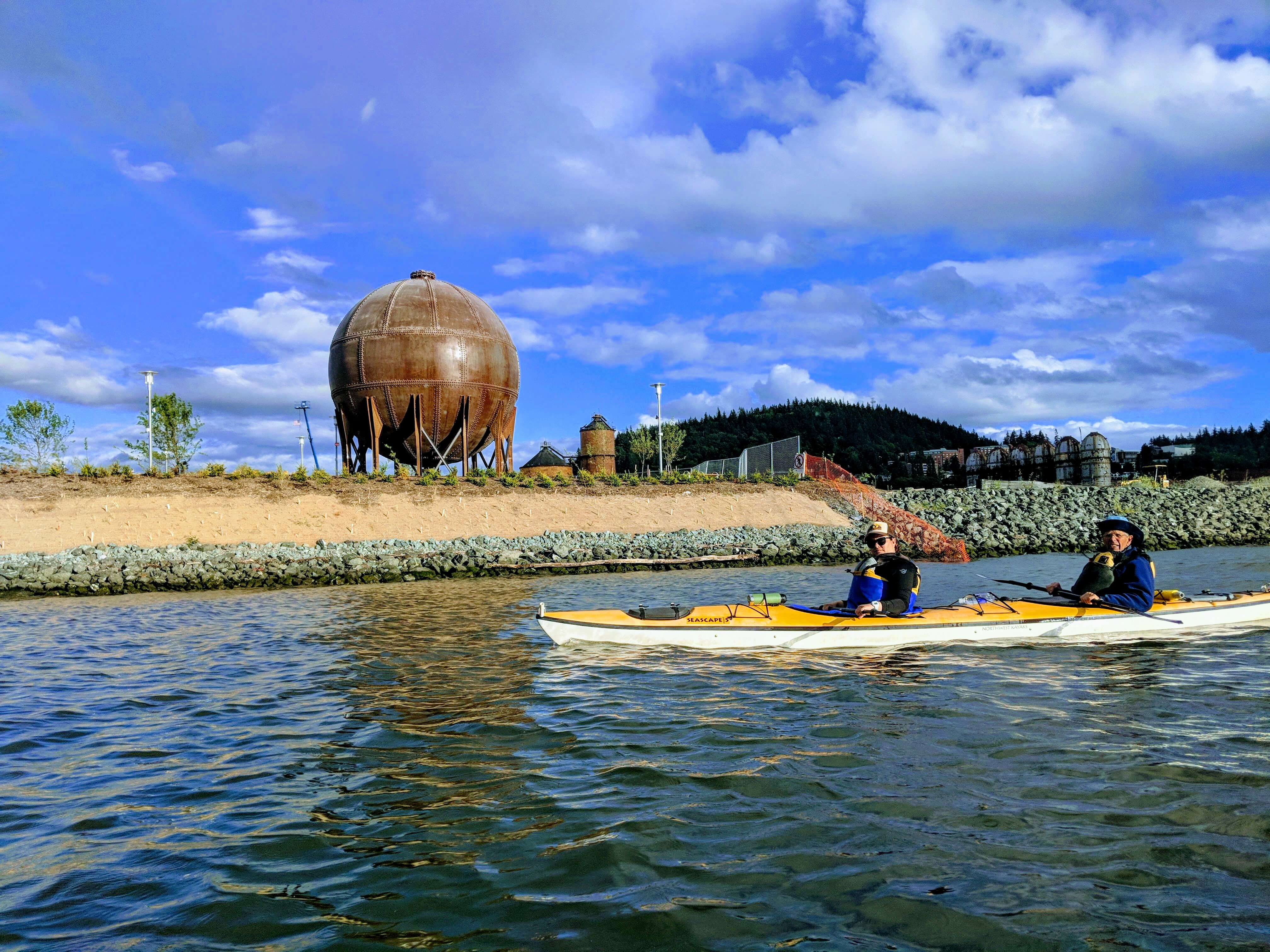 Two people in a kayak on the water. Behind them is a small beach and a large rusty metal sphere, a relic of old waterfront industry.