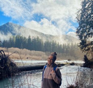 Sydney, a volunteer, stands smiling in front of a lake and trees with a partly cloudy blue sky.