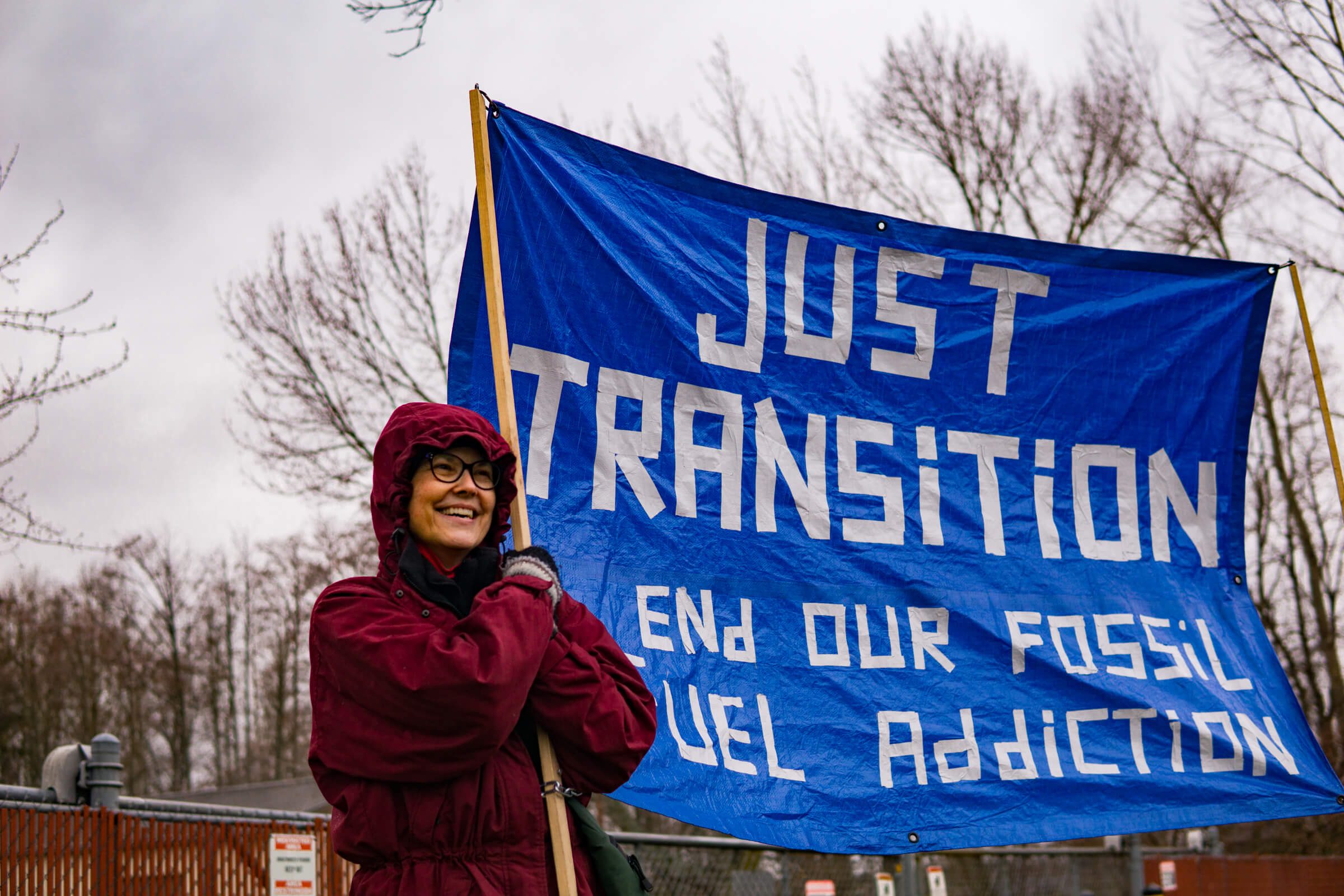 A person in a red coat smiling holds up a blue tarp with written words: "Just Transition, end our fossil fuel addiction"