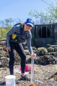 A person leaning over a turbidity tube full of water with an stormwater outfall in the background