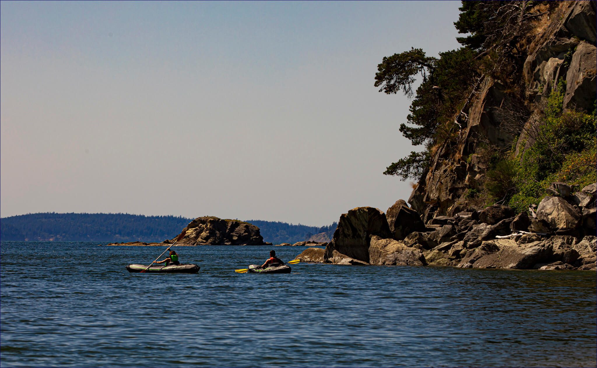 Two kayakers paddle near a sandstone bluff with trees overhanging it.